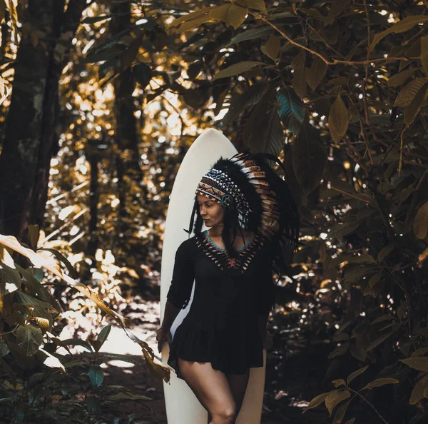 Sexy Maya-Häuptling mit Stammeskopfschmuck posiert im Wald vor einem Surfbrett — Stockfoto