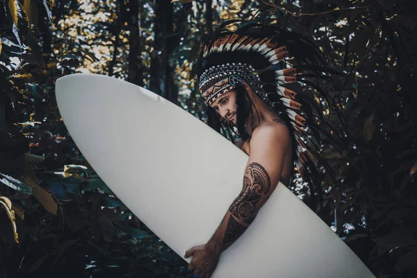 Inked and strong male model posing in a woods while holding a surfing board and wearing mayan feather hat — Stock Photo, Image