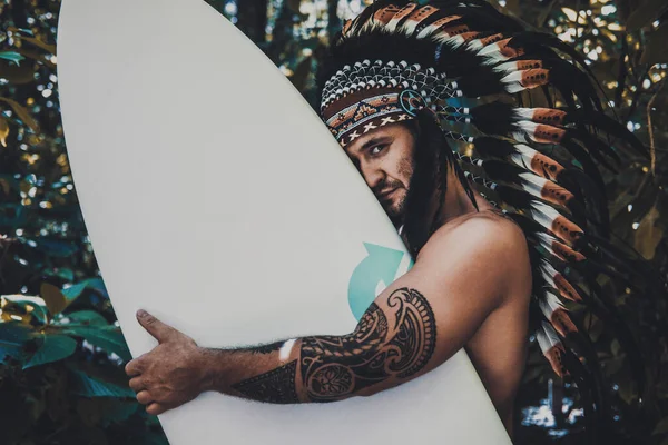 Mysterious and strong male with tattoos posing in a woods while holding a surfing board and wearing mayan feather hat