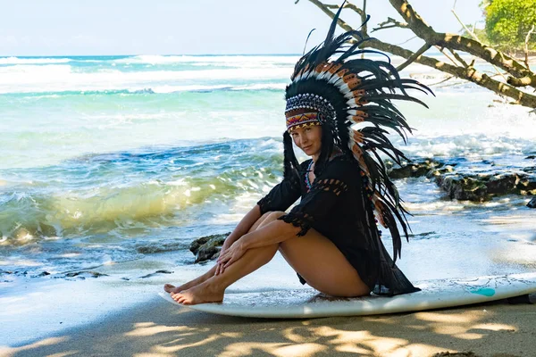 Half-nude female model sitting on a surfing board on a sandy beach in the sunshine and wearing ethnic indian feather headdress — Stock Photo, Image