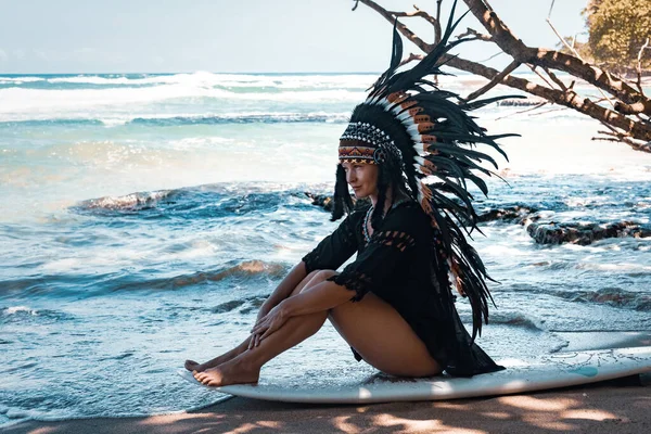 Attractive and fit woman posing while sitting on a surfing board and wearing mayan indian headwear on a beach near the ocean waves — Stock Photo, Image