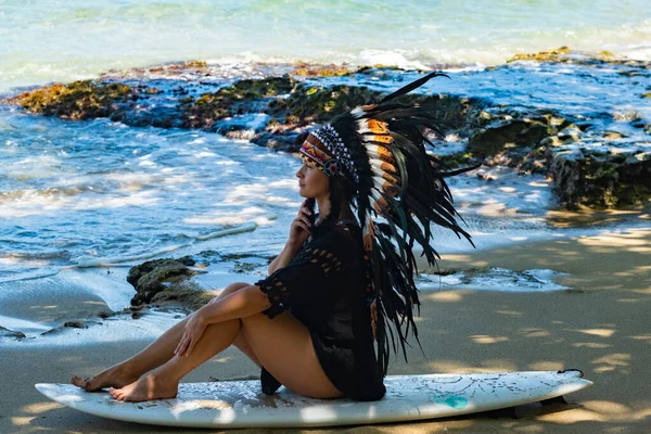 Attraktive und fitte Frau posiert auf einem Surfbrett und trägt Maya-Kopfbedeckungen an einem Strand in der Nähe der Meereswellen — Stockfoto