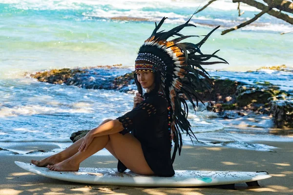 Halbnacktes weibliches Model sitzt auf einem Surfbrett an einem Sandstrand in der Sonne und trägt eine Kopfbedeckung aus ethnischen indischen Federn — Stockfoto