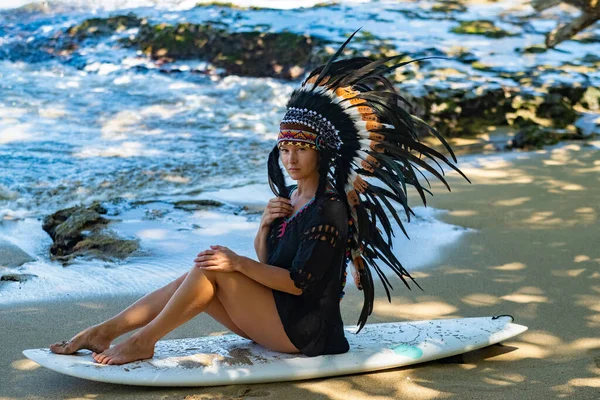 Sexy Maya-Häuptling trägt Stammeskopfschmuck, während er mit Surfbrett an einer Meeresküste sitzt — Stockfoto