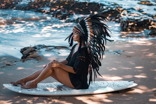 Sexy mayan female chief wearing tribal headdress while sitting on an ocean shore with surfing board