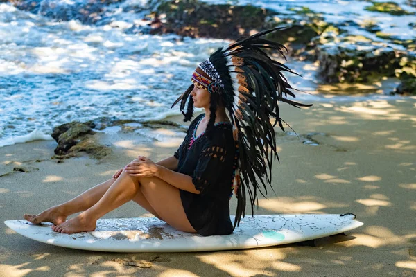 Attractive and fit woman posing while sitting on a surfing board and wearing mayan indian headwear on a beach near the ocean waves — Stock Photo, Image