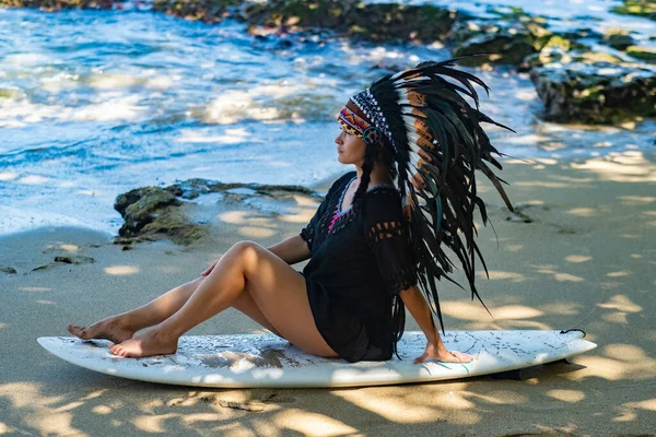 Half-nude female model sitting on a surfing board on a sandy beach in the sunshine and wearing ethnic indian feather headdress — Stock Photo, Image