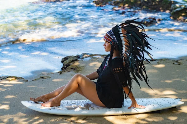 Half-nude female model sitting on a surfing board on a sandy beach in the sunshine and wearing ethnic indian feather headdress