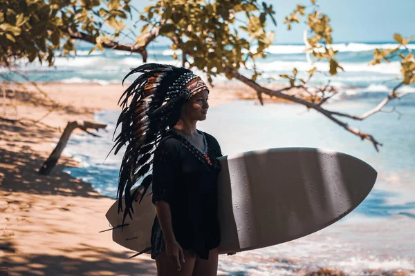 Attraktiv kvinna poserar i en skugga medan hon håller i en surfbräda och bär mayaindianska huvudbonader på en strand nära havsvågorna — Stockfoto