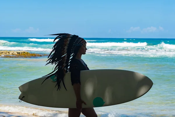 Half-nude female model holding a surfing board on a sandy beach in the shadow of sunshine and wearing ethnic indian feather headdress — Stock Photo, Image