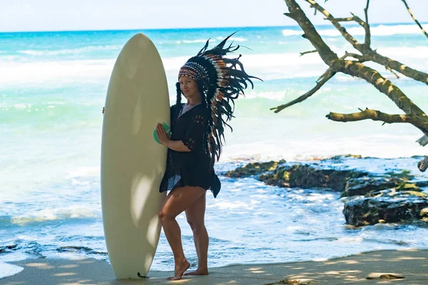 Half-nude female model holding a surfing board on a sandy beach in the shadow of sunshine and wearing ethnic indian feather headdress — Stock Photo, Image