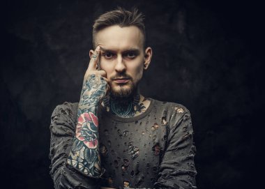 Koyu gri arka planda uzun kollu yırtık kazak giyen dövmeli erkek model.