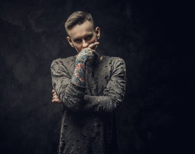 Koyu gri arka planda uzun kollu yırtık kazak giyen dövmeli erkek model.