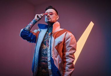 Neon bir stüdyoda yarı çıplak vücutlu, yarış ceketli ve güneş gözlüklü havalı bir erkek model poz veriyor..