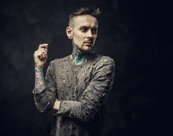 Inked young adult male wearing ripped jumper posing in a dark studio on a grey background — Stock Photo, Image