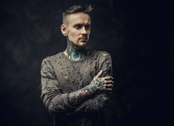 Inked young adult male wearing ripped jumper posing in a dark studio on a grey background — Stock Photo, Image