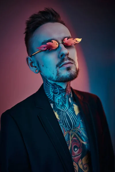 Cool tattoo artist male model posing in a neon studio with half-naked tattooed body, tuxedo and sunglasses. Vertical portrait close up