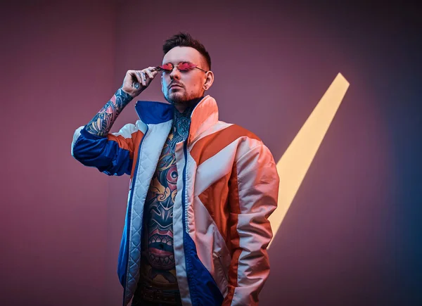 Modelo masculino fresco y rebelde posando en un estudio de neón con cuerpo tatuado semidesnudo, chaqueta de corredor y gafas de sol . — Foto de Stock