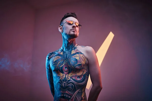 半裸のタトゥーボディとサングラスでネオンスタジオにポーズをとるタトゥーアーティストの男性モデル。縦の肖像 — ストック写真