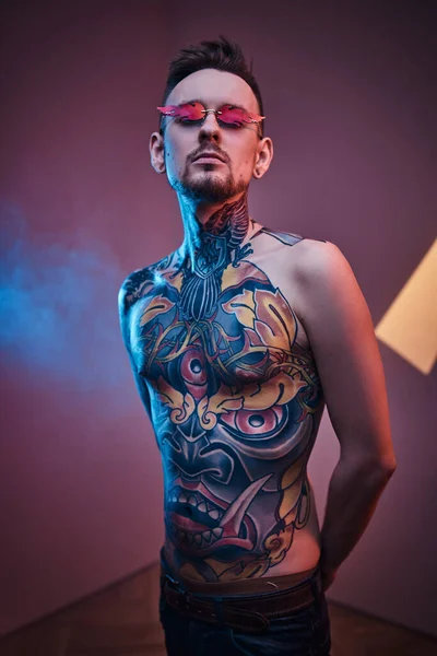 Tatuaje artista modelo masculino posando en un estudio de neón con cuerpo tatuado semidesnudo y gafas de sol. Retrato vertical — Foto de Stock