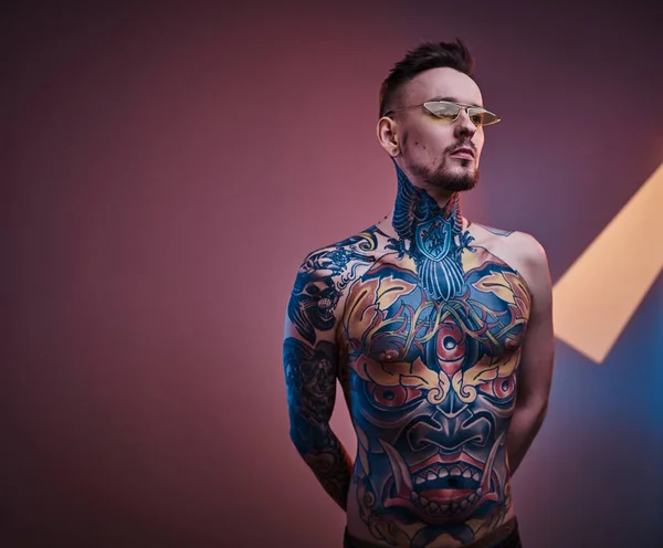 Modelo masculino tintado posando en un estudio de neón con cuerpo tatuado semidesnudo y gafas de sol. Retrato vertical — Foto de Stock