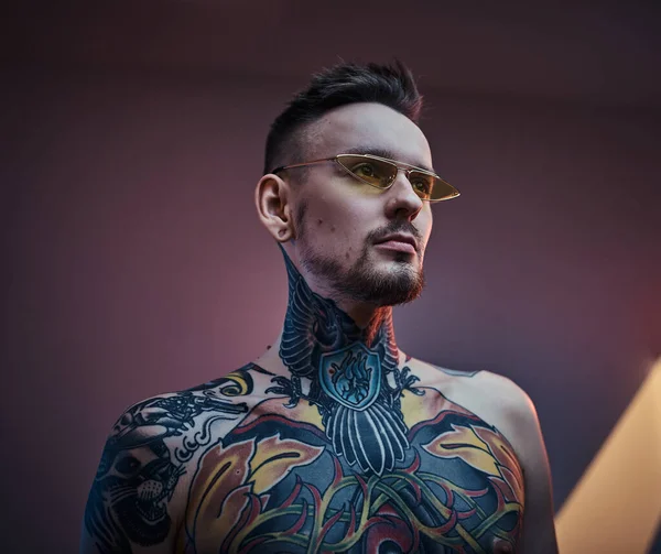 Tattoo artist male model posing in a neon studio with half-naked tattooed body and sunglasses. Vertical portrait