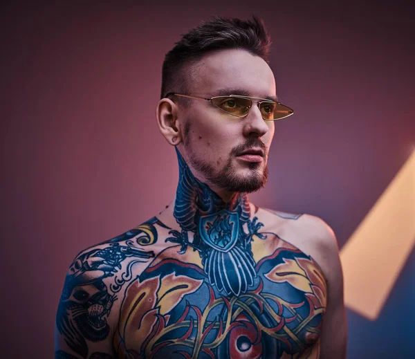 Tatuaje artista modelo masculino posando en un estudio de neón con cuerpo tatuado semidesnudo y gafas de sol. Retrato vertical — Foto de Stock