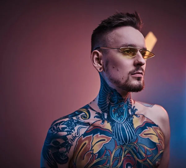 Tatuaje artista modelo masculino posando en un estudio de neón con cuerpo tatuado semidesnudo y gafas de sol. Retrato vertical — Foto de Stock