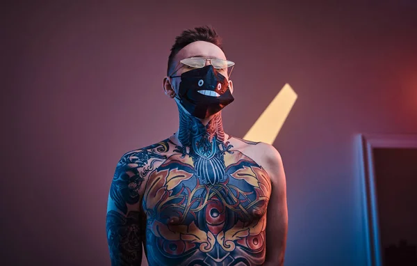 Infärgad manlig modell som poserar i en neonateljé med halvnaken tatuerad kropp, solglasögon och medicinsk mask. Vertikalt porträtt — Stockfoto