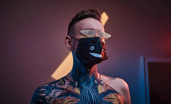 Крутой и чернильный мужчина-татуировщик позирует в неоновой студии с полуобнаженным телом, солнцезащитными очками и медицинской маской . — стоковое фото