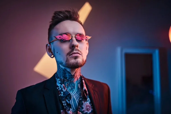 Tatueringsartist man modell poserar i en neon studio med halvnaken tatuerad kropp, smoking och solglasögon. — Stockfoto