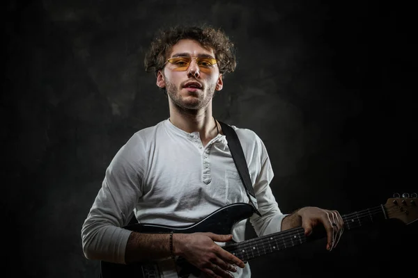 Portret van een knappe muzikant man met krullend haar poseren in de donkere studio met elektrische gitaar — Stockfoto