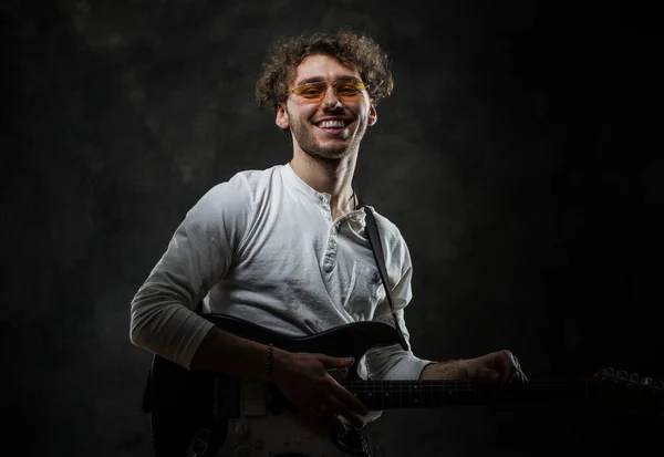 Musicien masculin attrayant souriant jouant une guitare électrique dans un studio sombre portant des vêtements décontractés et des lunettes de soleil — Photo