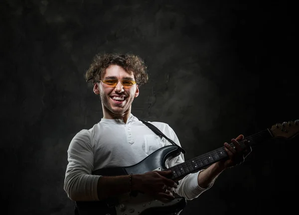 Улыбающийся привлекательный мужчина-музыкант, играющий на электрогитаре в темной студии в повседневной одежде и солнечных очках — стоковое фото