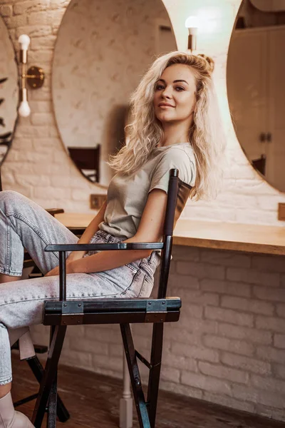 Mulher loira confiante com cabelo encaracolado sentado em uma cadeira para maquiagem no estúdio cheio de espelhos — Fotografia de Stock
