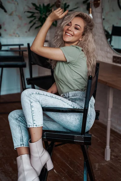 Με αυτοπεποίθηση ξανθιά γυναίκα με σγουρά μαλλιά κάθεται σε μια καρέκλα για μακιγιάζ στο στούντιο γεμάτο καθρέφτες — Φωτογραφία Αρχείου