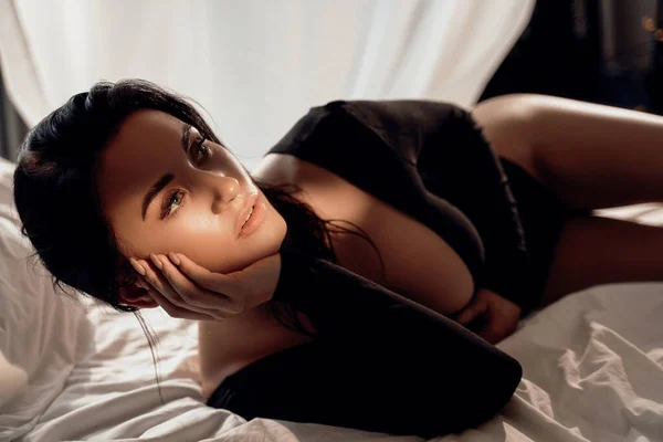 一个性感而性感的女人穿着黑色紧身衣躺在床单中间 手挽着她深棕色的头发的画像 — 图库照片