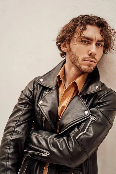 Daring and cool male model with curly hair posing in leather coat in a bright studio