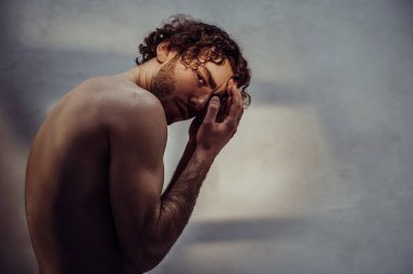 Çıplak ve şehvetli bir adam, kıvırcık saçlı, gri arka planda bir fotoşop için poz veriyor, kendini ışıktan koruyor ve elleriyle arkasını dönüyor.