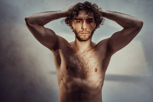 Shirtless young adult male model with curly hair posing in a dark studio, highlited with a sun, looking luscious and hot