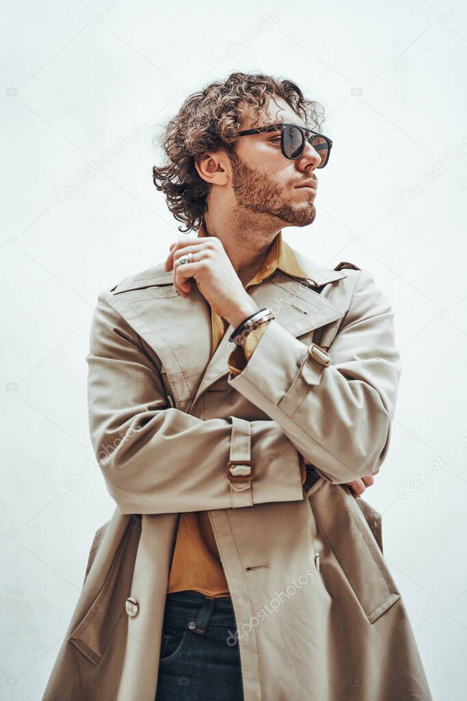 Curly and serious caucasian man standing in a bright studio on a white background, wearing casual and stylish trench coat and sunglasses