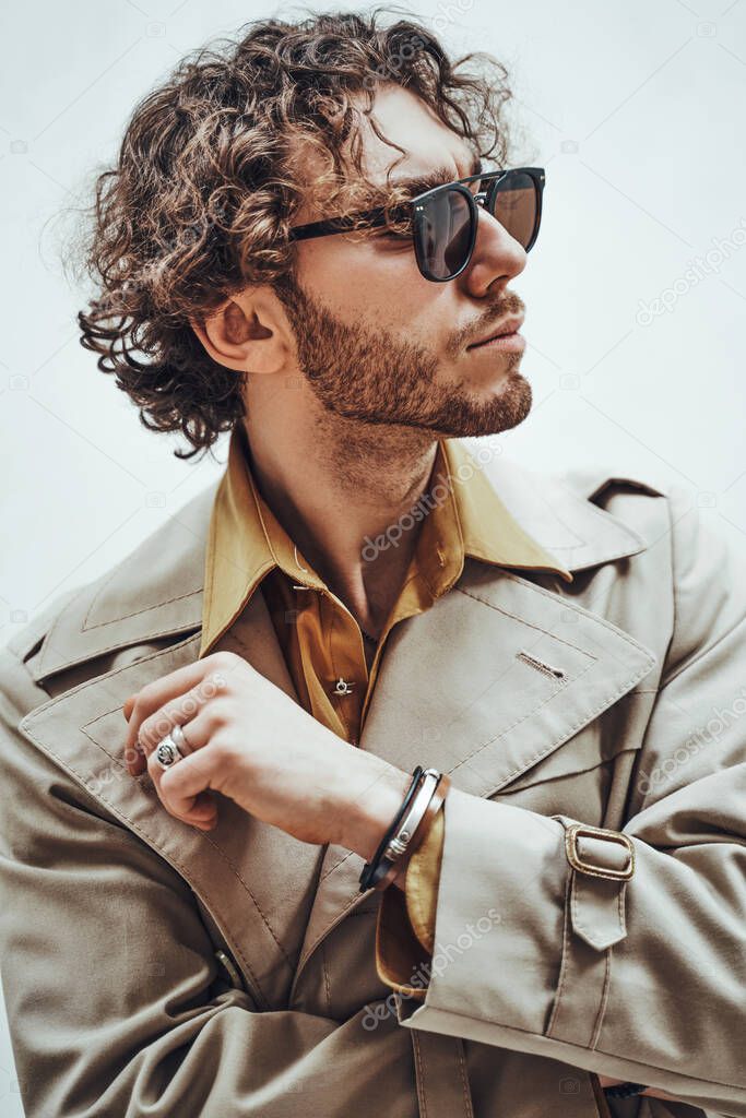 Thoughtful and handsome young man posing in a bright studio on a white background, looking interested while wearing trench coat and glasses