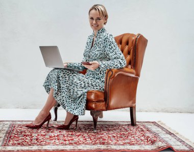 Parlak bir stüdyoda deri bir sandalyede oturan, mavi bir elbise giyen ve elinde gülümseyerek dizüstü bilgisayar tutan zarif bir iş kadını.