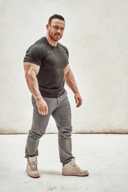 Atletik adam beyaz bir arka planda izole edilmiş, günlük kıyafetler giyiyor ve kabartmalı vücudunu gösteriyor, ciddi görünüyor.