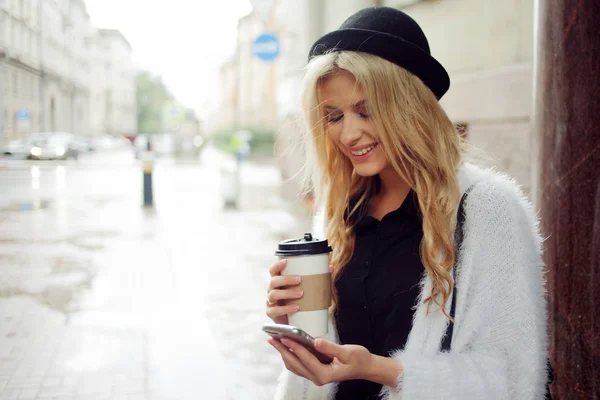 Vrolijke vrouw in de straat drinken koffie in de ochtend en haar smartphone gebruiken. — Stockfoto