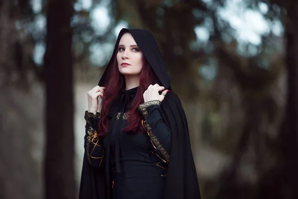 Joven mujer hermosa y misteriosa en el bosque, en capa negra con capucha, imagen de elfo bosque o bruja — Foto de Stock
