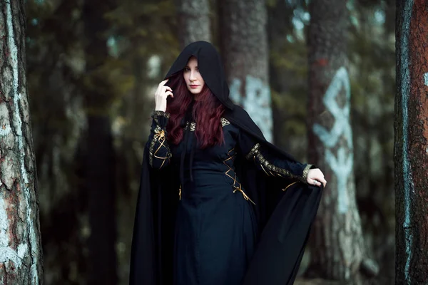 Jovem mulher bonita e misteriosa na floresta, em manto preto com capuz, imagem de elfo da floresta ou bruxa — Fotografia de Stock