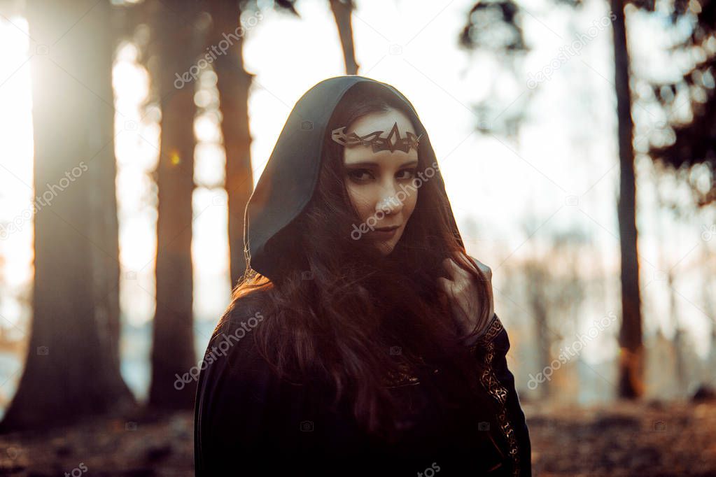 Young beautiful and mysterious woman in woods, in black cloak with hood, image of forest elf or witch