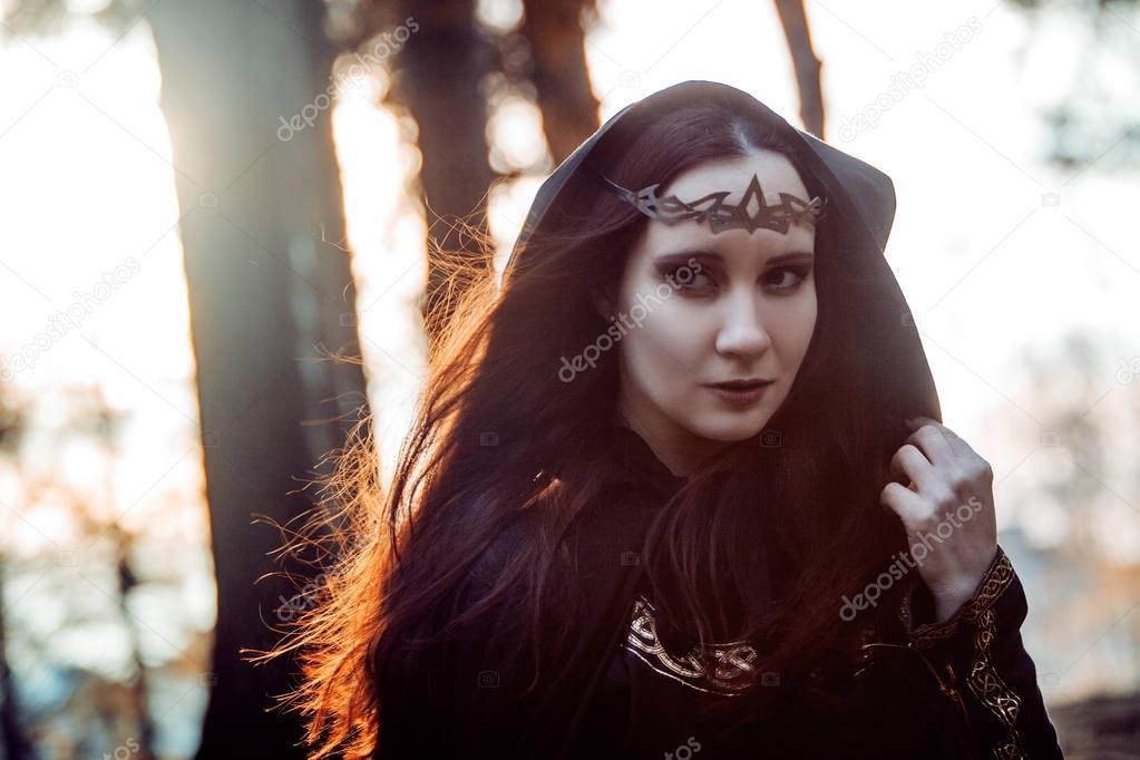 Young beautiful and mysterious woman in woods, in black cloak with hood, image of forest elf or witch