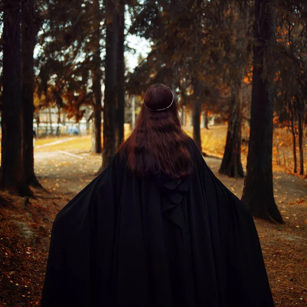 Jovem mulher bonita e misteriosa na floresta, em manto preto com capuz, imagem de elfo da floresta ou bruxa, de volta — Fotografia de Stock